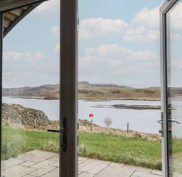 A view through open glass doors showing a patio leading to grass and water with hills in the background at Sula in Cullipool Isle Of Luing