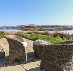 An outdoor patio with two wicker chairs and a table with two mugs overlooking a lake and grassy area at Sula Cullipool Isle Of Luing