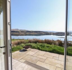 View through open glass doors onto a stone patio with grass and a body of water with hills in the background at Scarba in Cullipool Isle Of Luing