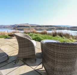 An outdoor patio with wicker chairs and a small table overlooking water and hills at Torsa Cullipool Isle Of Luing
