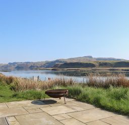 A patio with a fire pit overlooking a body of water and hills at Torsa in Cullipool Isle Of Luing
