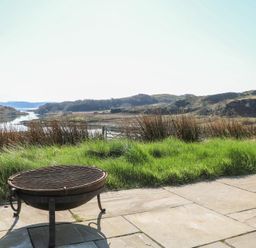 A fire pit on a stone patio with grassy area and hills in the background at Torsa Cullipool Isle Of Luing