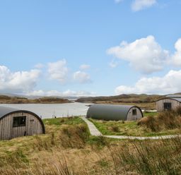 Two semi cylindrical buildings with wood siding near water with hills in background at Fladda Cullipool Isle Of Luing