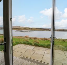 A view of a grassy landscape and a body of water through open glass doors at Fladda in Cullipool Isle Of Luing