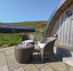 An outdoor seating area with two wicker chairs and a table with two mugs in front of a wooden pod at Losal in Cullipool Isle Of Luing
