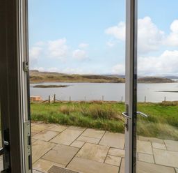 View through open glass doors onto a stone patio and grassy area overlooking a loch with hills in the distance at Losal Cullipool Isle Of Luing