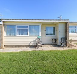 A single story building with windows chairs and tables on a stone patio in front of a lawn at Chalet 37 in Hemsby