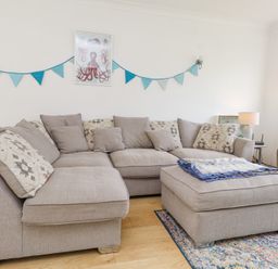 A living room with a beige sectional sofa, matching ottoman, blue bunting on wall, and an octopus picture at 3 Glyn Y Mor in Llanbedrog