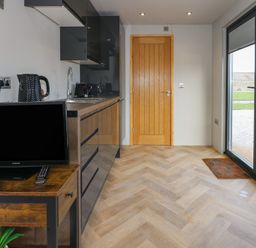 A kitchen area with black cabinets a wooden TV stand with a TV kettle and toaster wooden door and glass sliding doors showing a garden outside at Caban in Dothan near Gwalchmai
