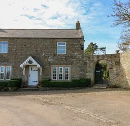A stone cottage with white windows and door beside an arched stone gate at Pagecroft Cottage in Haydon Bridge