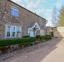 Exterior stone wall of a house with windows a white door with a small porch cobblestone pathway and trimmed hedges at Pagecroft Cottage in Haydon Bridge