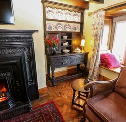 A room with a fireplace wooden cabinet dining plates vase with flowers upholstered chair and window seat at Pagecroft Cottage in Haydon Bridge
