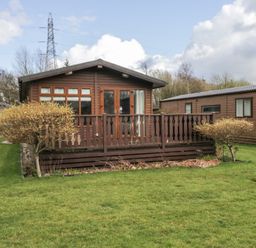 A wooden cabin with a front porch and railing surrounded by grass and small trees at Gressingham View Lodge Dock Acres near Carnforth