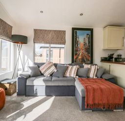 A living room with a gray sectional sofa with cushions and a red throw blanket a floor lamp an orange ottoman a side table and windows at Gressingham View Lodge in Dock Acres near Carnforth
