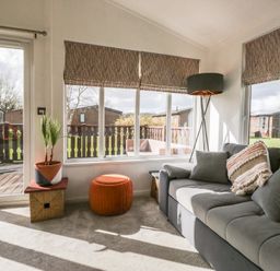 A living room with a gray sofa and patterned cushions near large windows with blinds a floor lamp a plant in a pot and an orange ottoman at Gressingham View Lodge Dock Acres near Carnforth