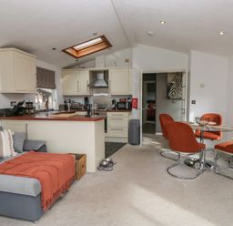 A living area with grey sofa and orange chairs around a glass table near a kitchen with cream cabinets at Gressingham View Lodge Dock Acres near Carnforth