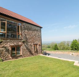 Stone cottage with wooden window frames and flower pots on a grassy lawn with trees and countryside in the background at Newlands Barn in Wellington