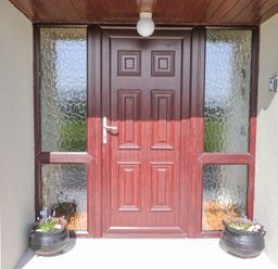 A wooden front door with textured glass panels on either side and flower pots in black containers at Teach Ards in Kilgarvan County Kerry