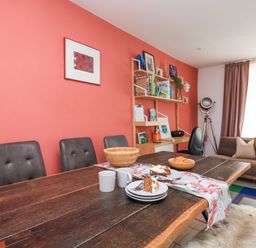 A dining room with a wooden table with food and mugs and chairs against a coral wall with bookshelves and a sofa at Joymead in Malborough