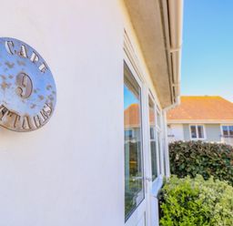 A white exterior wall with a round metal sign reading cape 9 cottages next to glass doors and bushes at Cape A9 Norton Near Freshwater