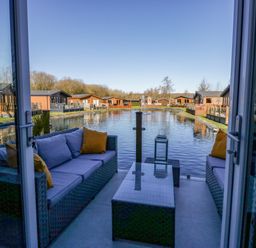 A patio with outdoor sofas and a table overlooking a water canal with lodges on both sides at Coniston View Lodge in Warton near Carnforth