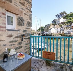 A small outdoor area with a table holding a bottle of prosecco two glasses and a plate of scones beside a blue railing overlooking boats and houses at Harbour View in Polperro