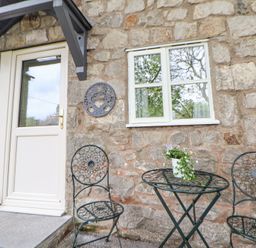 An outdoor seating area with two metal chairs and a metal table with a flower pot in front of a stone wall with a door and window at Cuddfan at Swallow Cottage in Rhydtalog near Coedpoeth