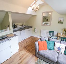 A small kitchen area with white cabinets and appliances next to a living area with a grey sofa and colorful cushions at Cuddfan at Swallow Cottage in Rhydtalog near Coedpoeth