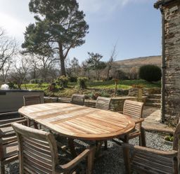 An outdoor wooden dining table with chairs on a gravel patio next to a stone wall with trees and a hill in the background at White Haywood Farmhouse & Barn in Craswall