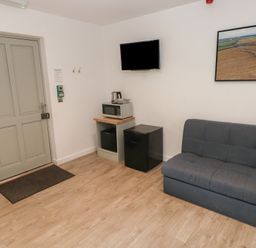 A room with a grey door a small microwave stand a black mini fridge a wall-mounted TV a grey couch and framed wall art at Sycamore in Cowbridge