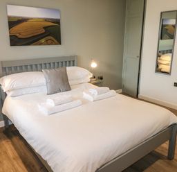 A bedroom with a double bed white bedding towels on bed two bedside tables with lamps wall mirror and landscape picture at Alder in Cowbridge
