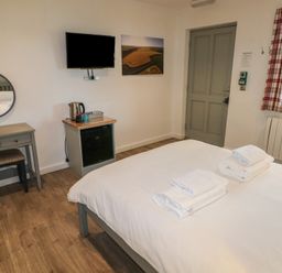 A bedroom with a bed with towels on it a desk with a round mirror a television on the wall a small fridge and a window with plaid curtains at Cherry in Cowbridge