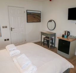 A bedroom with a bed and towels on it a small desk with a mirror a mini fridge with a kettle and cups and a wall-mounted television at Cherry in Cowbridge