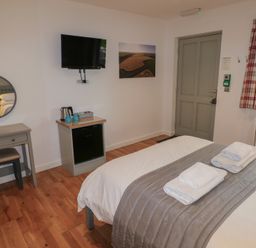 A bedroom with a bed with folded towels a small desk with a stool a wall-mounted TV a kettle on a cabinet and a window with plaid curtains at Chestnut in Cowbridge