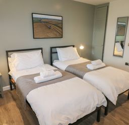 A bedroom with two single beds with white bedding and gray runners a bedside table with a lamp and a wall mirror at Oak in Cowbridge
