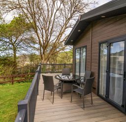 An outdoor deck with a round glass table and four chairs next to a building with large glass doors at 16 Manleigh Park in Combe Martin
