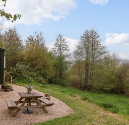 An outdoor area with a wooden picnic table and flowers by a green hut and grassy trees at Oak Shepherd Hut in Monkton Wyld near Charmouth