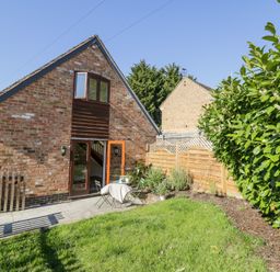 A brick house with an open door leading to a patio with a table and chairs next to a garden with green grass and shrubs at Little Barn in Dodwell near Stratford-Upon-Avon
