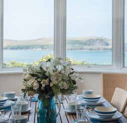 A dining table set with blue and white dishes and glasses with a floral centerpiece by windows overlooking the sea at Splits in Daymer Bay