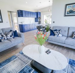 A living room with two grey sofas a white oval coffee table with flowers a blue and grey rug and a kitchen with blue cabinets at Number 5 Villa - The View in Derrybeg County Donegal