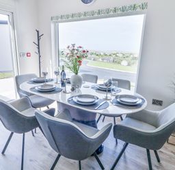 A dining area with a white oval table set for six with grey chairs and a vase of flowers by a large window at Number 5 Villa The View in Derrybeg County Donegal
