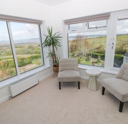 A sitting area with two beige chairs a small round table and large windows overlooking a garden and countryside at Burry House in Llanmorlais
