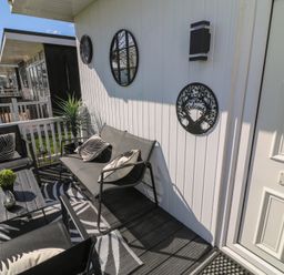 A small outdoor seating area with black chairs, a table with a plant, decorative wall hangings, and a white door at 56 Cherry Park Chalet in Chapel St Leonards