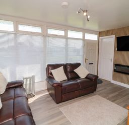 A living room with leather sofas cushions a TV on the wall an electric fireplace and a window with blinds at 56 Cherry Park Chalet in Chapel St Leonards