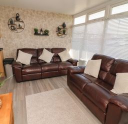 A living room with two brown leather sofas with white cushions and wall shelves with plants at 56 Cherry Park Chalet in Chapel St Leonards