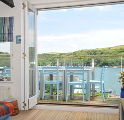 A room with an open glass door leading to a balcony with blue chairs and a table overlooking a river and green hills at 5 Esplanade in Fowey