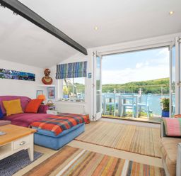 A living room with a purple sofa, colorful cushions, striped rugs, a coffee table, and an open door leading to a balcony with blue chairs overlooking water at 5 Esplanade in Fowey