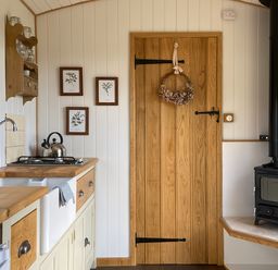 A kitchen with a wooden countertop a white sink a kettle on a stove a wooden door with a wreath and a black wood stove in the corner at Shepherd Hut 1 in Northchapel