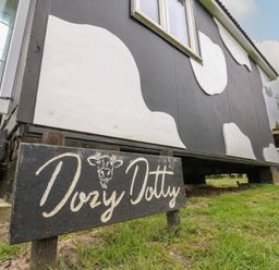 A small building painted with black and white cow patterns and a wooden sign reading Dozy Dotty outside on grass at Dozy Dotty (Cow Shed 4) Easton near Campsea Ashe