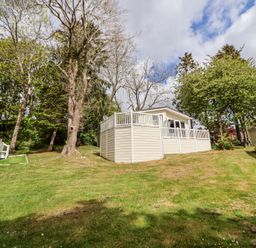 A mobile home with a white deck on a grassy hill surrounded by trees at 155 Woodland Hall Lyons in Ruthin